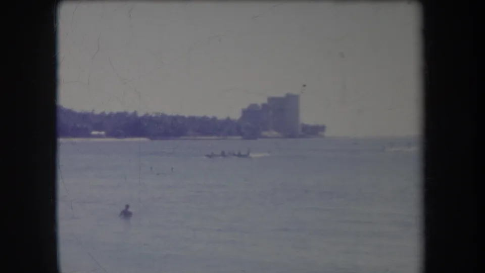1969 Hawaii 8mm frame showing calm coastal water, a distant palm-lined shore, a large building, small craft, and a swimmer.