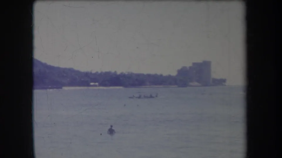 1969 Hawaii coastal view with a distant shore, palm trees, large building, small boat, and swimmer on calm water.