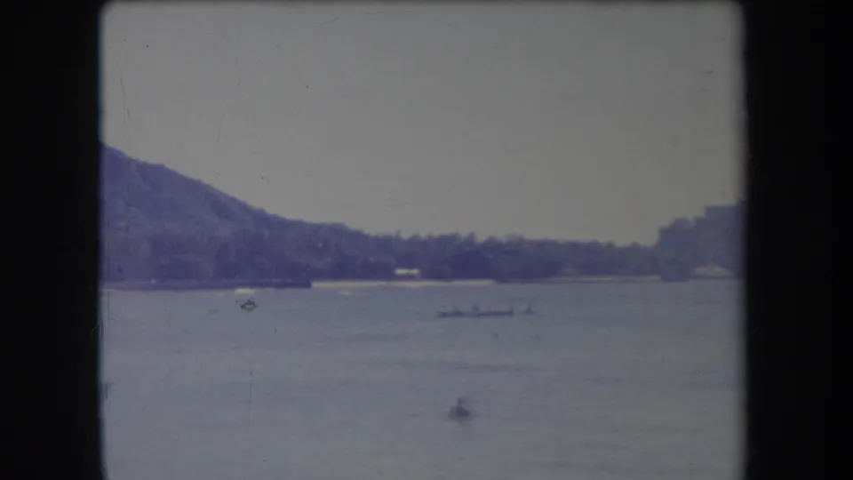 1969 Hawaii 8mm frame showing a mountain slope, palm-lined shore, calm sea, small craft, and a distant swimmer.