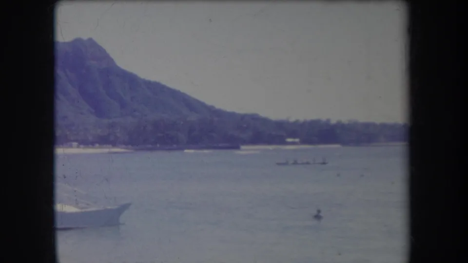 1969 Hawaii waterfront frame with a mountain backdrop, palm-lined shore, outrigger canoe, swimmer, and partial boat bow.