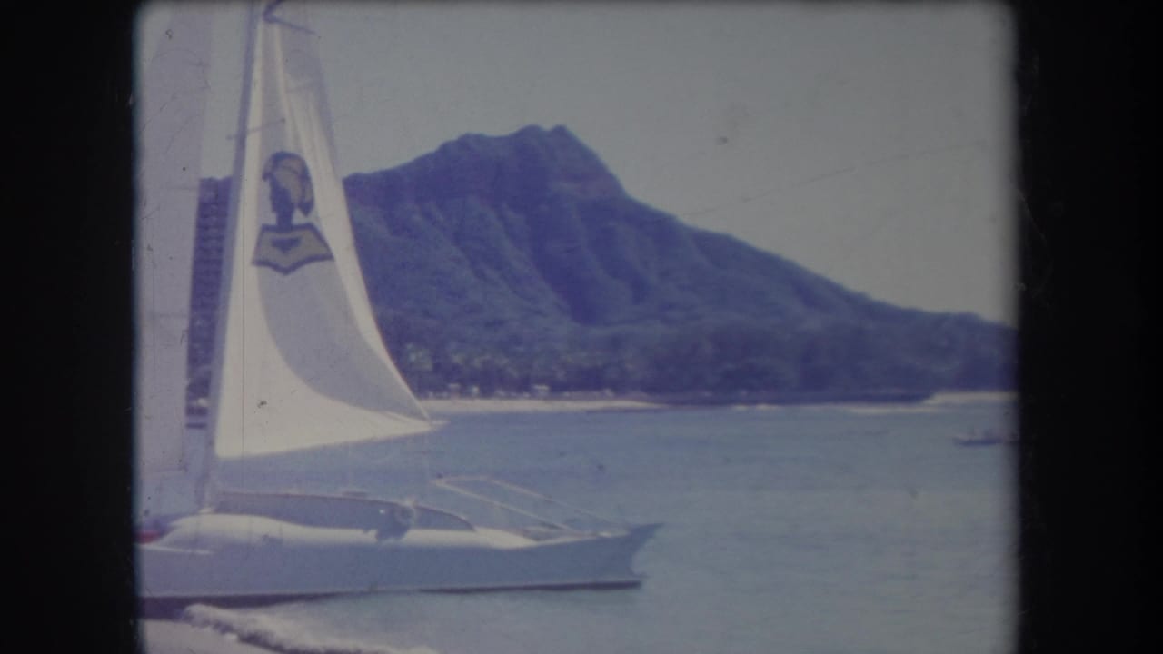 White sailboat with a marked sail passes in front of Diamond Head across calm water.