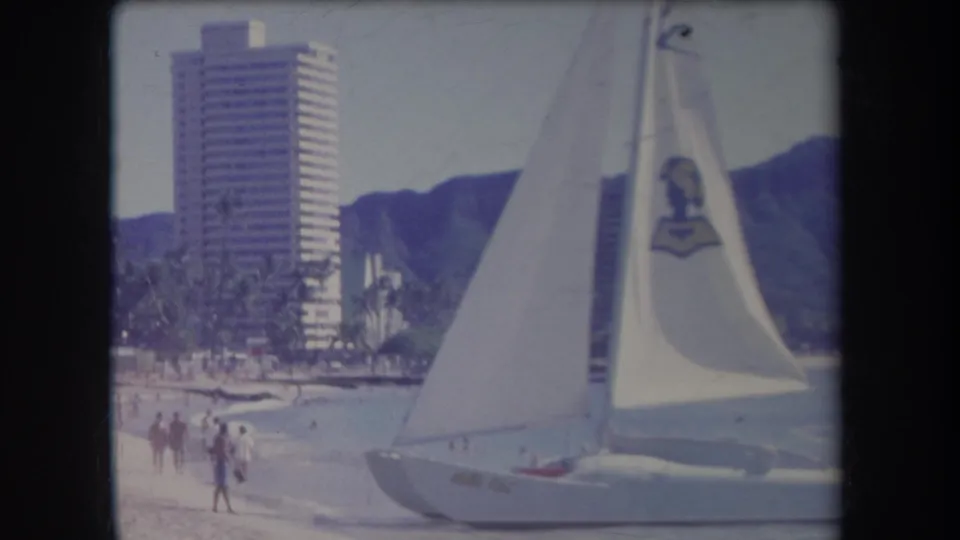 Beachgoers walk along the sand as a white sailboat passes near shore, with a tall resort building and Diamond Head in the background.