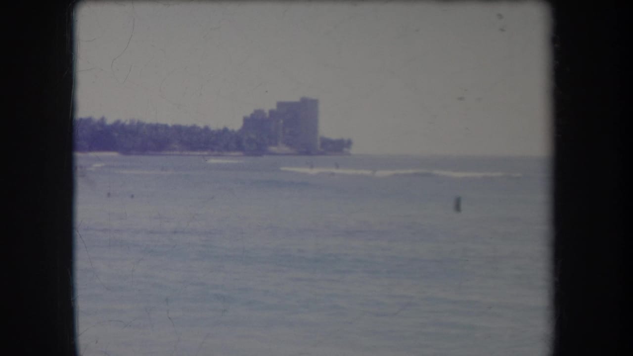 1969 Hawaii 8mm frame showing rough ocean water and a distant tropical shoreline with palm trees and high-rise buildings.