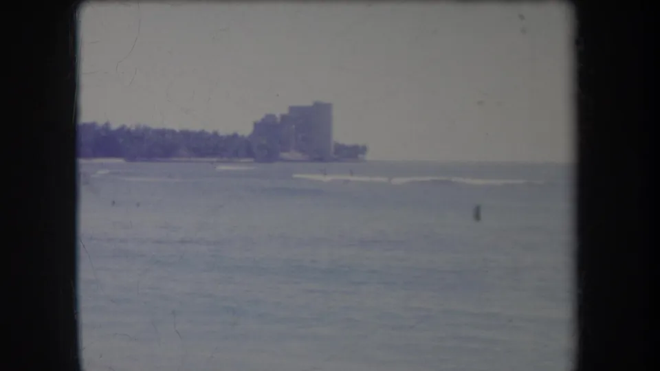 1969 Hawaii 8mm frame showing rough ocean water and a distant tropical shoreline with palm trees and high-rise buildings.