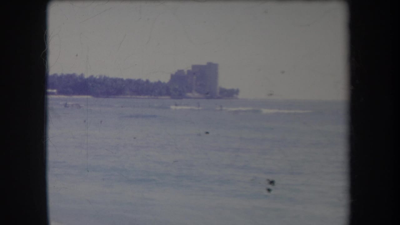 Distant Hawaii coastline in a 1969 home-movie frame, with surf lines, palms, and high-rise buildings softened by aged film color.