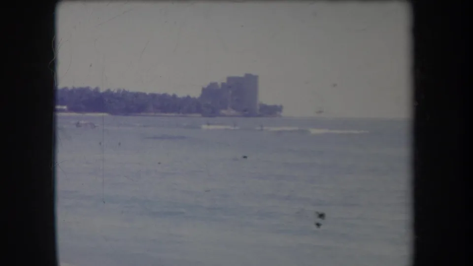 Distant Hawaii coastline in a 1969 home-movie frame, with surf lines, palms, and high-rise buildings softened by aged film color.