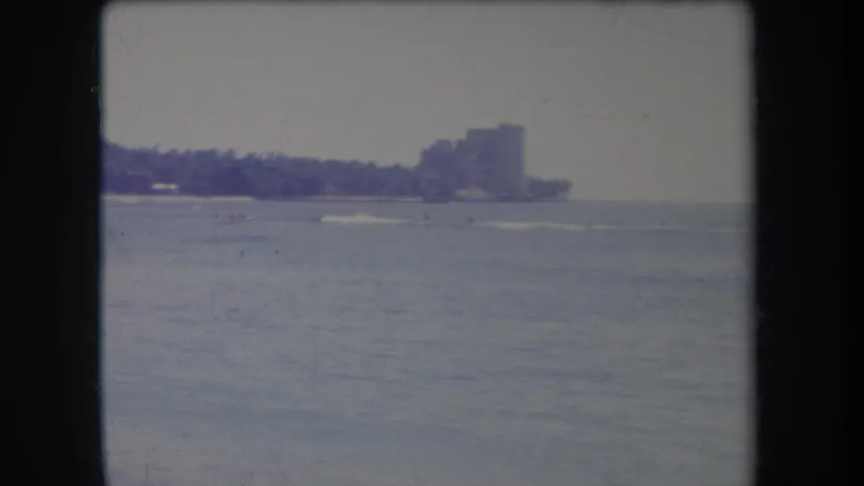 Vintage 8mm view of choppy water facing a distant Hawaii shoreline with palms and clustered high-rise buildings.