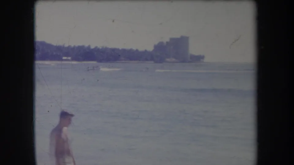 1969 Hawaii beach home-movie frame with rough ocean, distant buildings, palm-lined coast, and a partial passerby at the bottom edge.