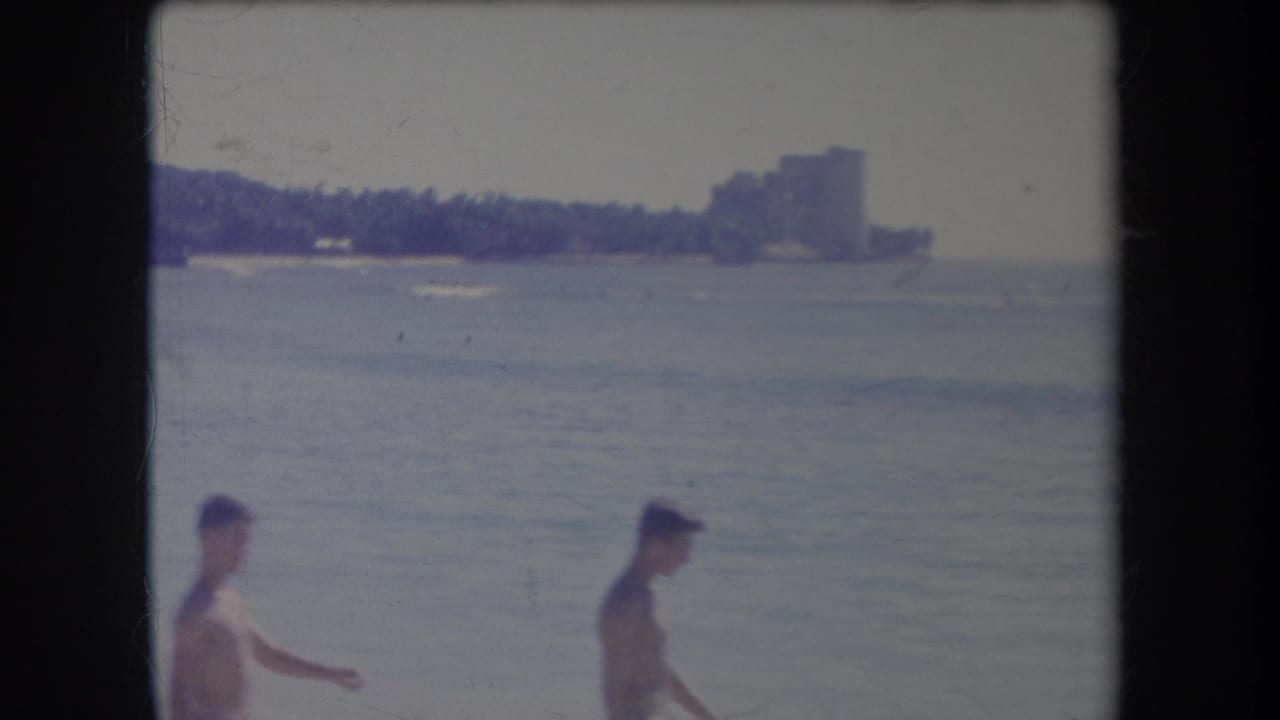 1969 Hawaii 8mm frame showing beachgoers near the water, a calm bay, and a distant shoreline structure.