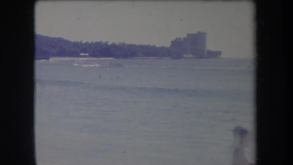 1969 Hawaii beach frame with a person near the waterline and small distant boats crossing the bay.