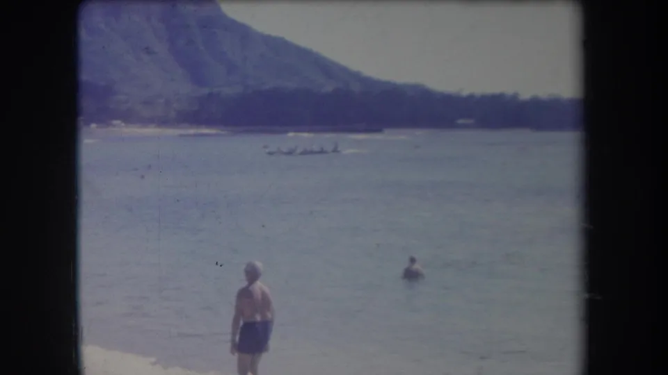 1969 Hawaii 8mm frame showing a man in blue shorts on the beach, a swimmer, and a longboat beyond shallow water.