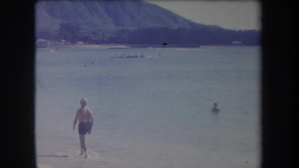 1969 Hawaii beach frame with a man in blue shorts near the water, a swimmer, distant longboat, and mountain backdrop.