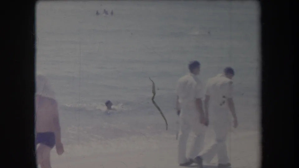1969 Hawaii shoreline frame showing two men in white clothing walking past ocean swimmers.