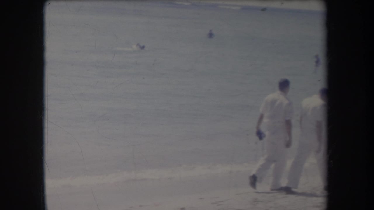 Two men in white walk along a sandy beach beside calm surf.