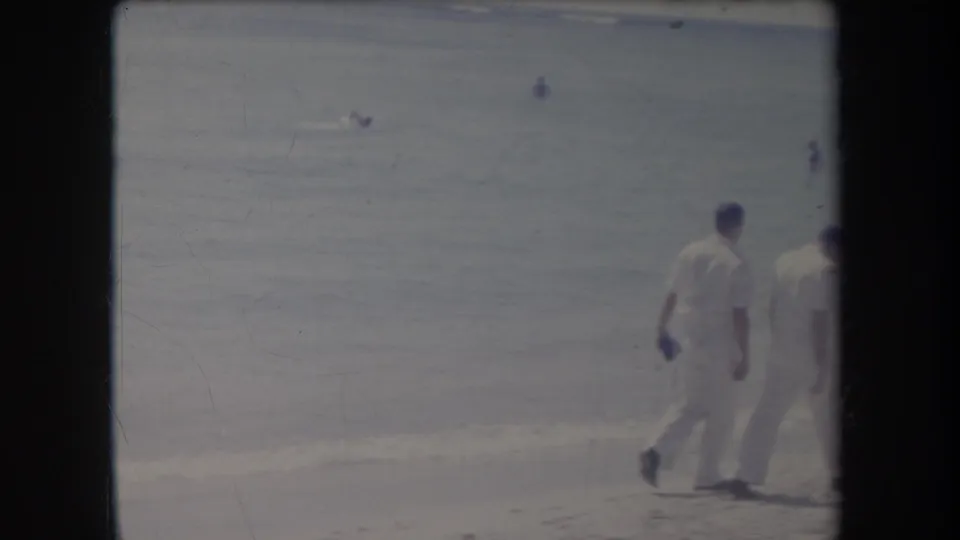 Two men in white clothing walk along a Hawaii beach in 1969 home-movie footage, with ocean and distant swimmers behind them.