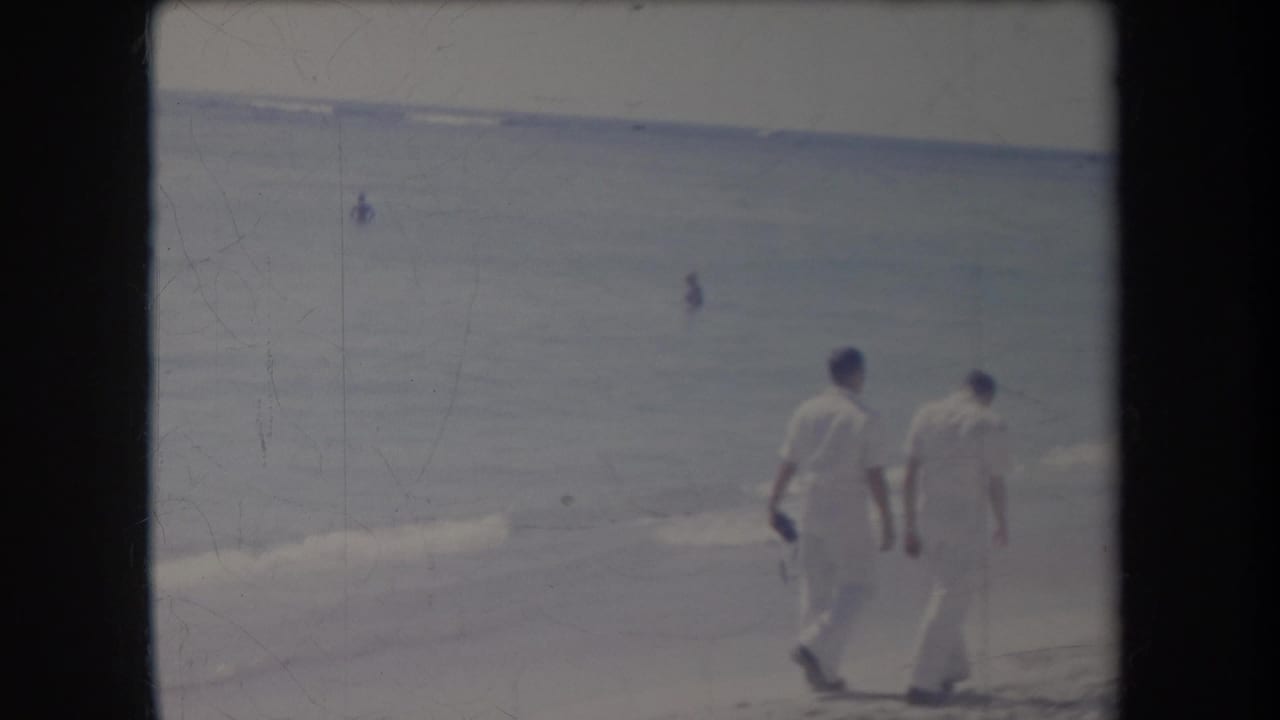 In a 1969 Hawaii beach scene, two men in white walk beside the surf while small figures appear in the water.