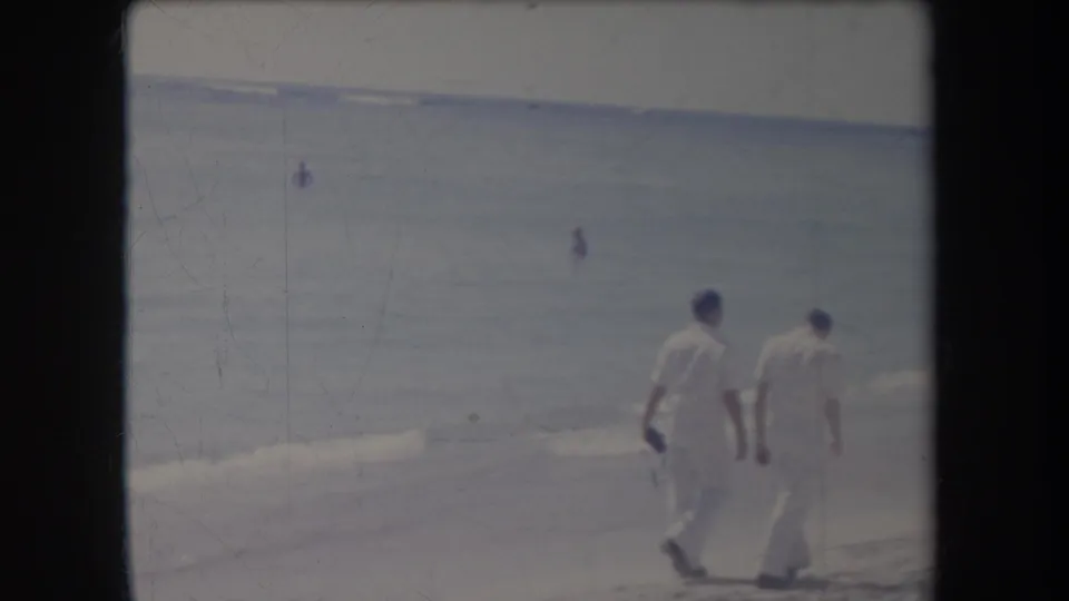 In a 1969 Hawaii beach scene, two men in white walk beside the surf while small figures appear in the water.