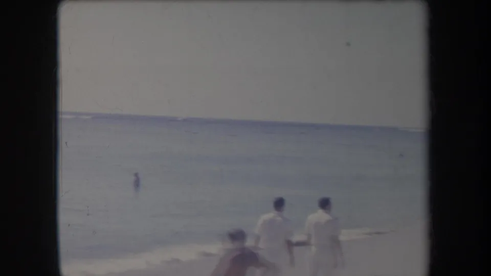 Two men in white walk down a Hawaii shoreline while a blurred beachgoer passes in the lower foreground of the 1969 footage.