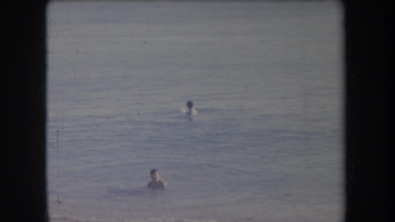 1969 Hawaii 8mm frame showing two bathers separated in calm ocean water, with faded color and black film borders.