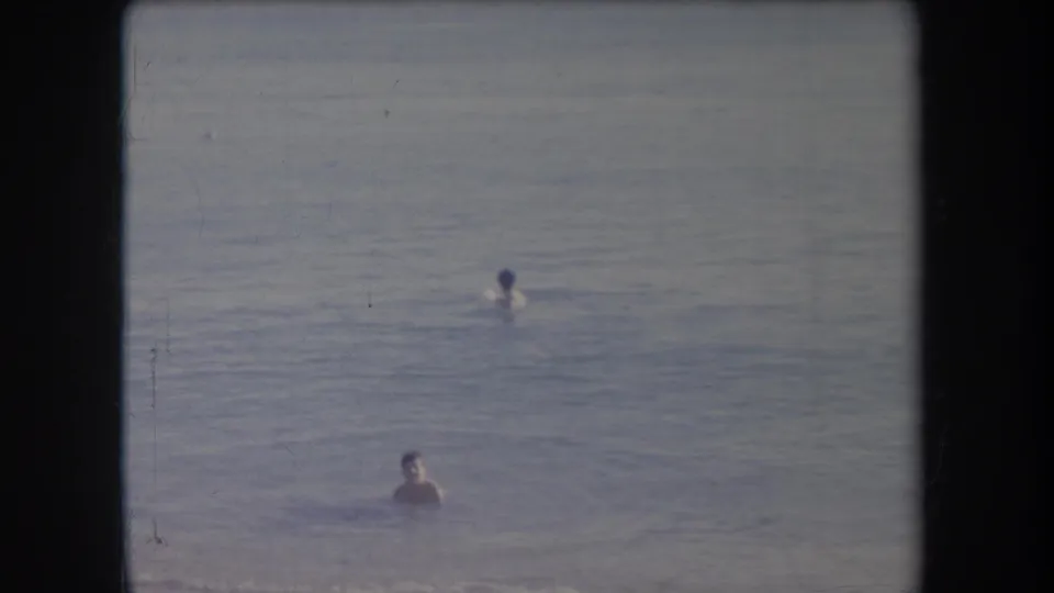 1969 Hawaii 8mm frame showing two bathers separated in calm ocean water, with faded color and black film borders.