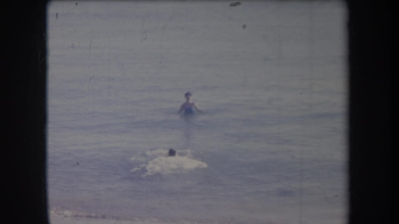 A person stands in shallow ocean water while another swimmer moves toward them with splashing.