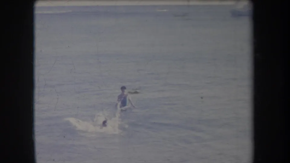 A smaller swimmer creates a splash while nearing a standing bather in the surf, framed by soft 8mm home-movie texture from Hawaii in 1969.