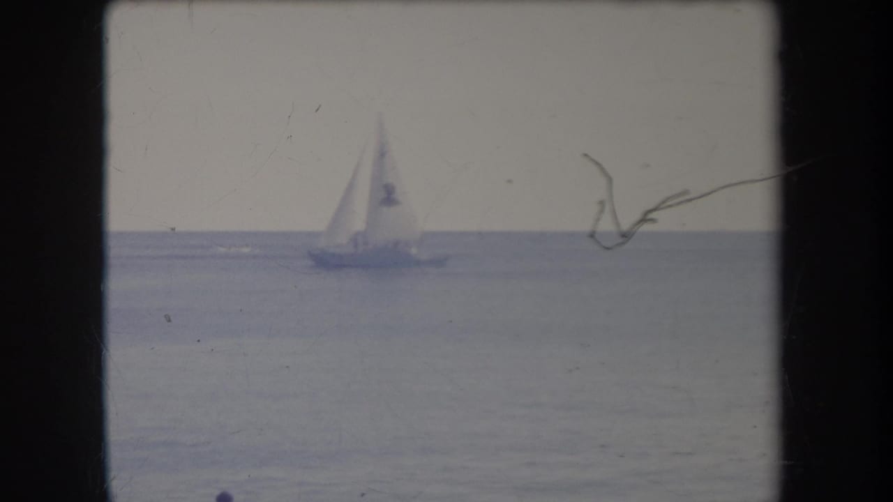 1969 Hawaii 8mm frame showing a distant sailboat crossing calm open water under a pale sky, with film scratches and gate edges visible.