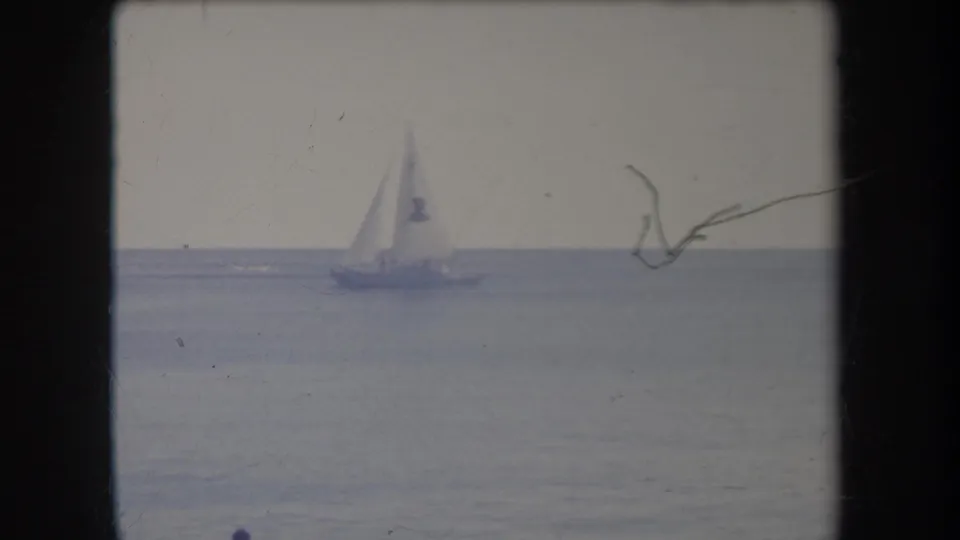 1969 Hawaii 8mm frame showing a distant sailboat crossing calm open water under a pale sky, with film scratches and gate edges visible.