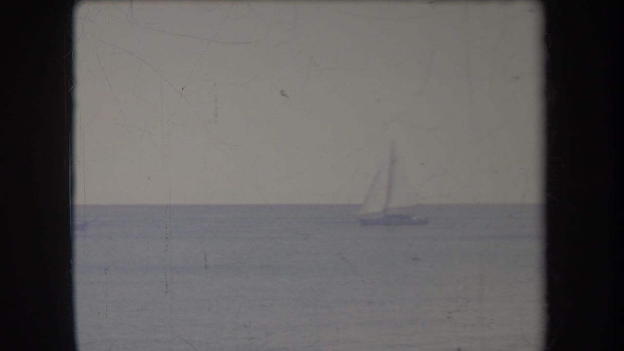 Small sailboat near the horizon on calm sea beneath a washed pale sky.