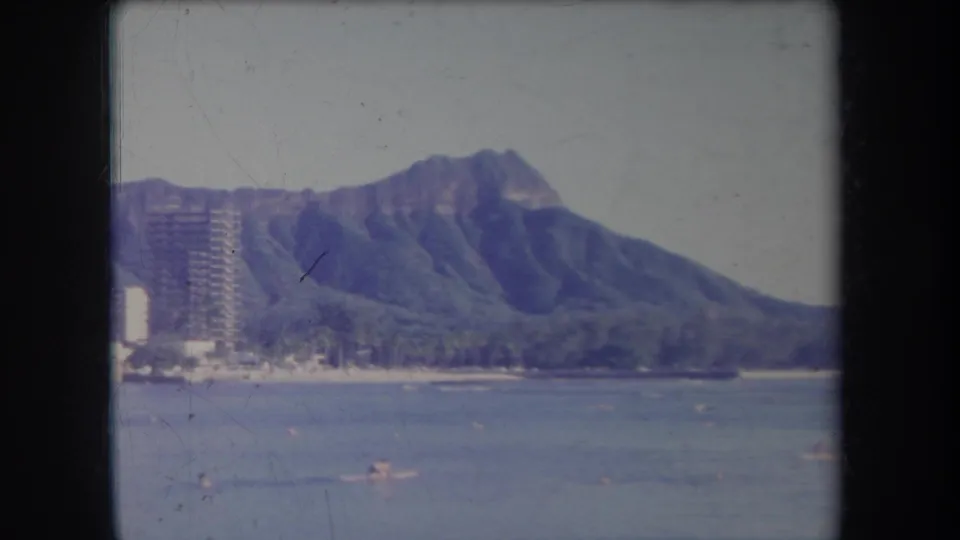 1969 Hawaii 8mm frame showing Diamond Head beyond blue water, distant swimmers, and shoreline high-rises at the edge of the beach.