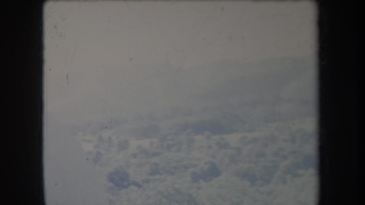 1969 8mm aerial view showing pale clouds and distant snow-covered mountain terrain, softened by haze and film wear.