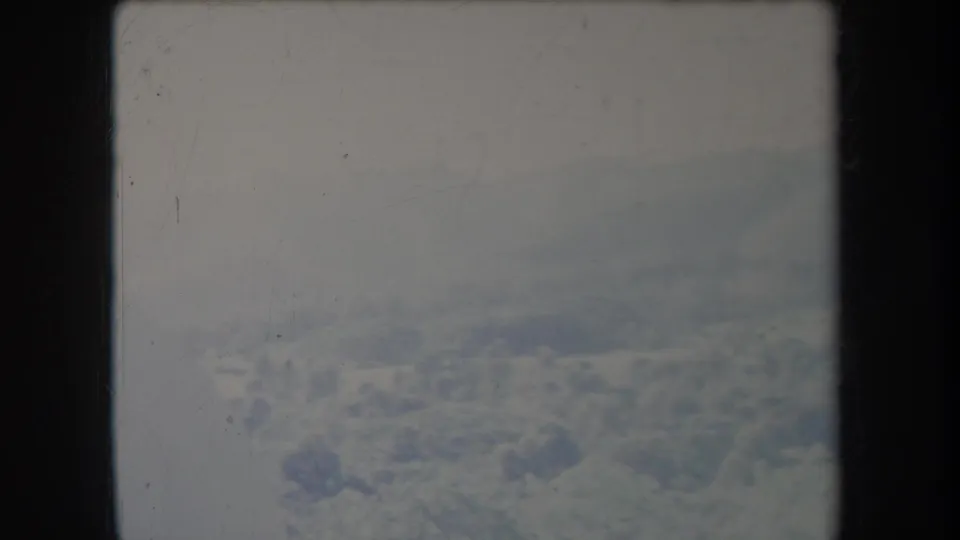1969 8mm aerial view showing pale clouds and distant snow-covered mountain terrain, softened by haze and film wear.