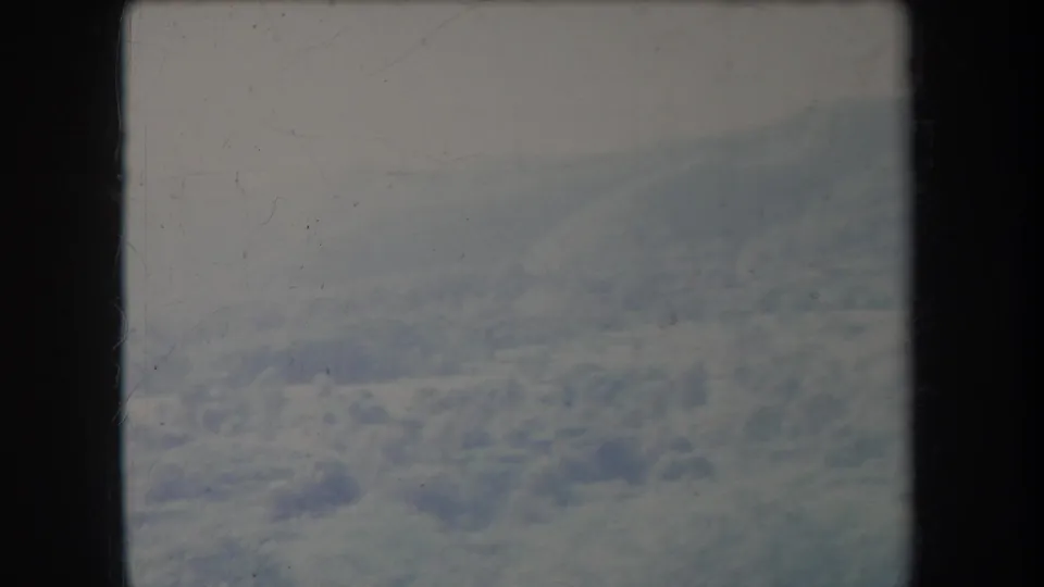 Vintage 8mm aerial frame with cloud cover below and a sloping snow-covered mountain ridge fading into haze.