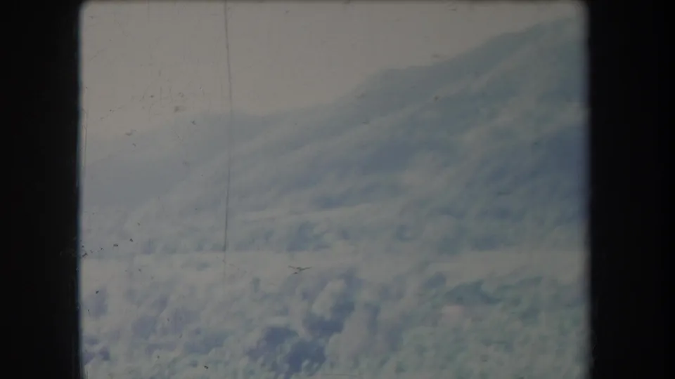 Vintage 8mm aerial frame of a snowy mountain slope and clouds below, seen through haze with visible film scratches.