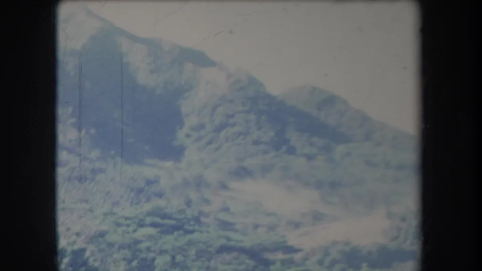 1969 Hawaii home-movie still with a blue-green mountain ridge, hazy sky, soft focus, and dark 8mm frame edges.
