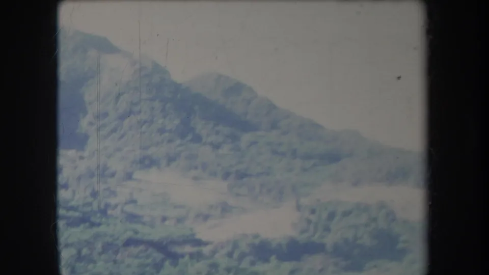 1969 Hawaii 8mm frame showing a sloping forested mountain and light-toned valley area, softened by haze and film fade.