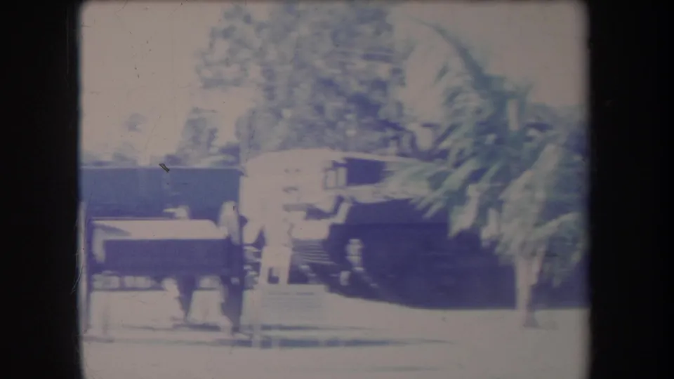 1969 Hawaii frame showing a palm-lined outdoor exhibit area with benches, barriers, and a dark display object in bright sun.