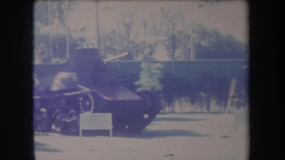 1969 Hawaii frame showing a small tank on display outdoors, with palms, fencing, grass, and a low sign nearby.