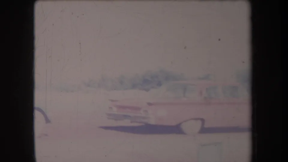 1969 Hawaii home-movie frame of a red mid-century sedan parked on a pale driveway before dense trees.