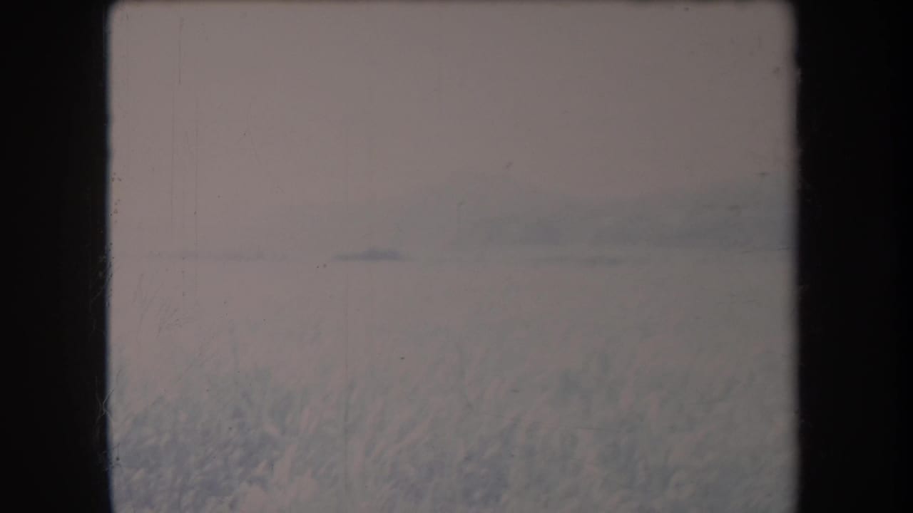Hazy grassy valley with distant hills and pale sky, seen within an 8mm film frame.