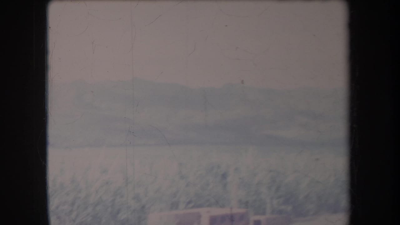 1969 Hawaii 8mm frame showing a pale crop field, hazy mountains, and a small indistinct structure at the bottom of the view.