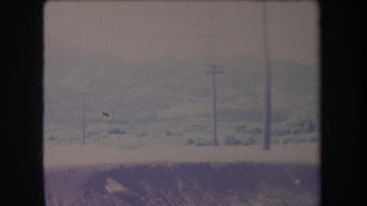 Hazy 1969 Hawaii landscape with distant rolling hills, utility poles, power lines, and a pale roadside strip in a faded 8mm frame.