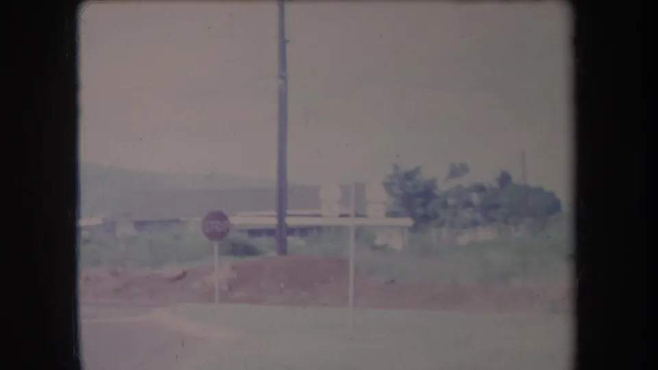 Vintage Hawaii 8mm frame showing a roadside stop sign near vegetation, a utility pole, and low buildings.