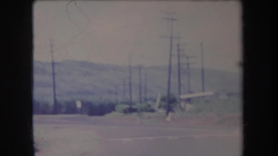 1969 Hawaii roadside frame with utility poles, wires, low vegetation, and a hazy mountain in the distance.