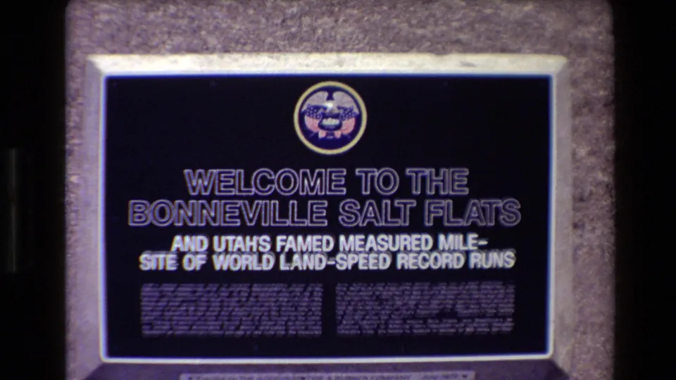 1980s 8mm frame of the Bonneville Salt Flats welcome sign in Utah, with white lettering on a dark plaque and a circular emblem above.