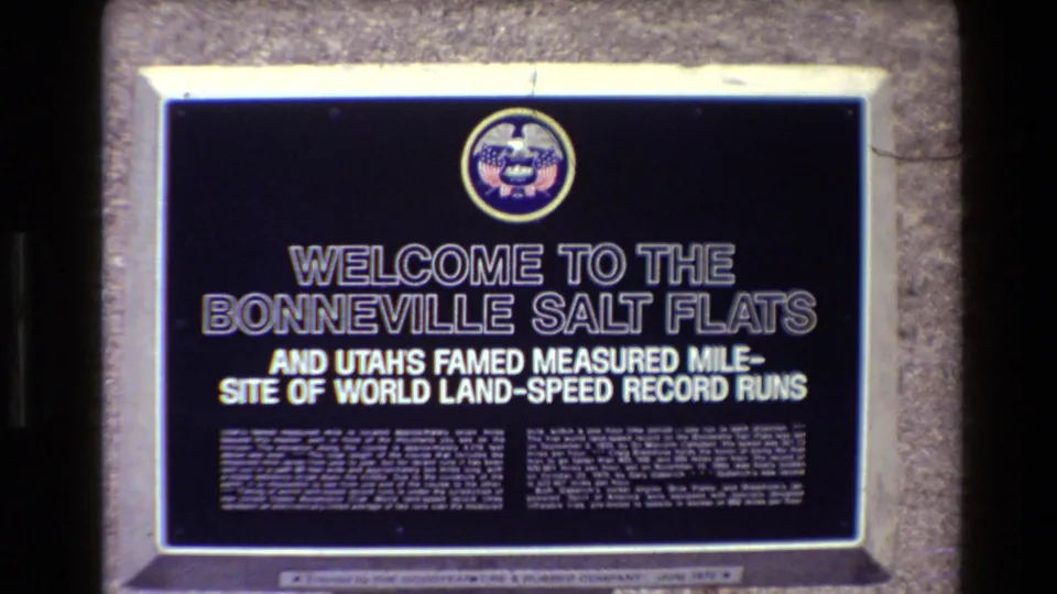 Visitor-shot 8mm view of the Bonneville Salt Flats welcome sign, with the dark plaque framed by a pale stone or concrete border.