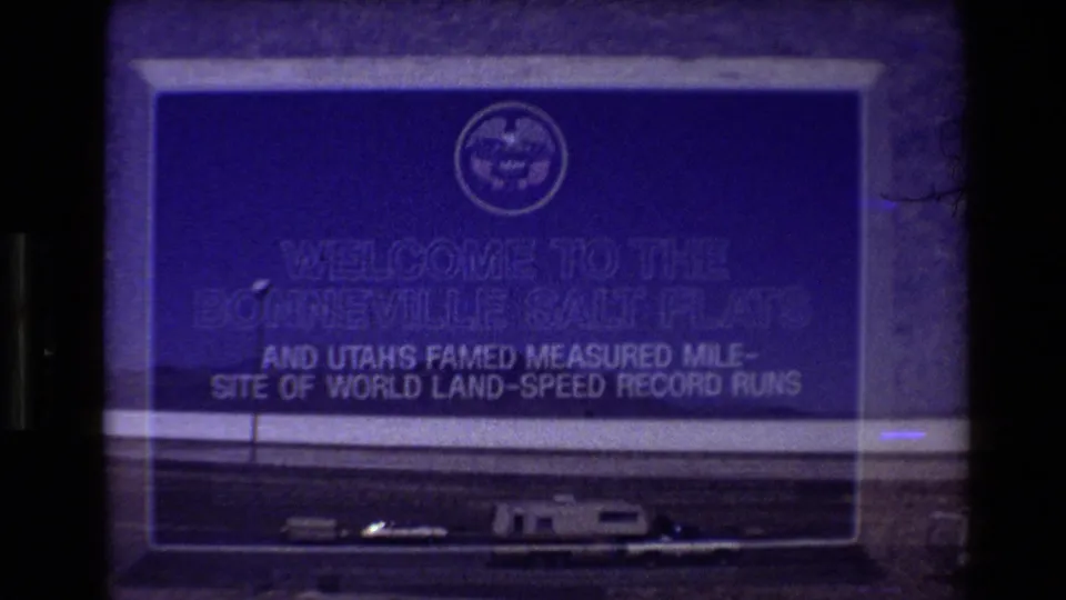 1980 8mm frame showing a Bonneville Salt Flats welcome sign above parked vehicles at the edge of the flats.