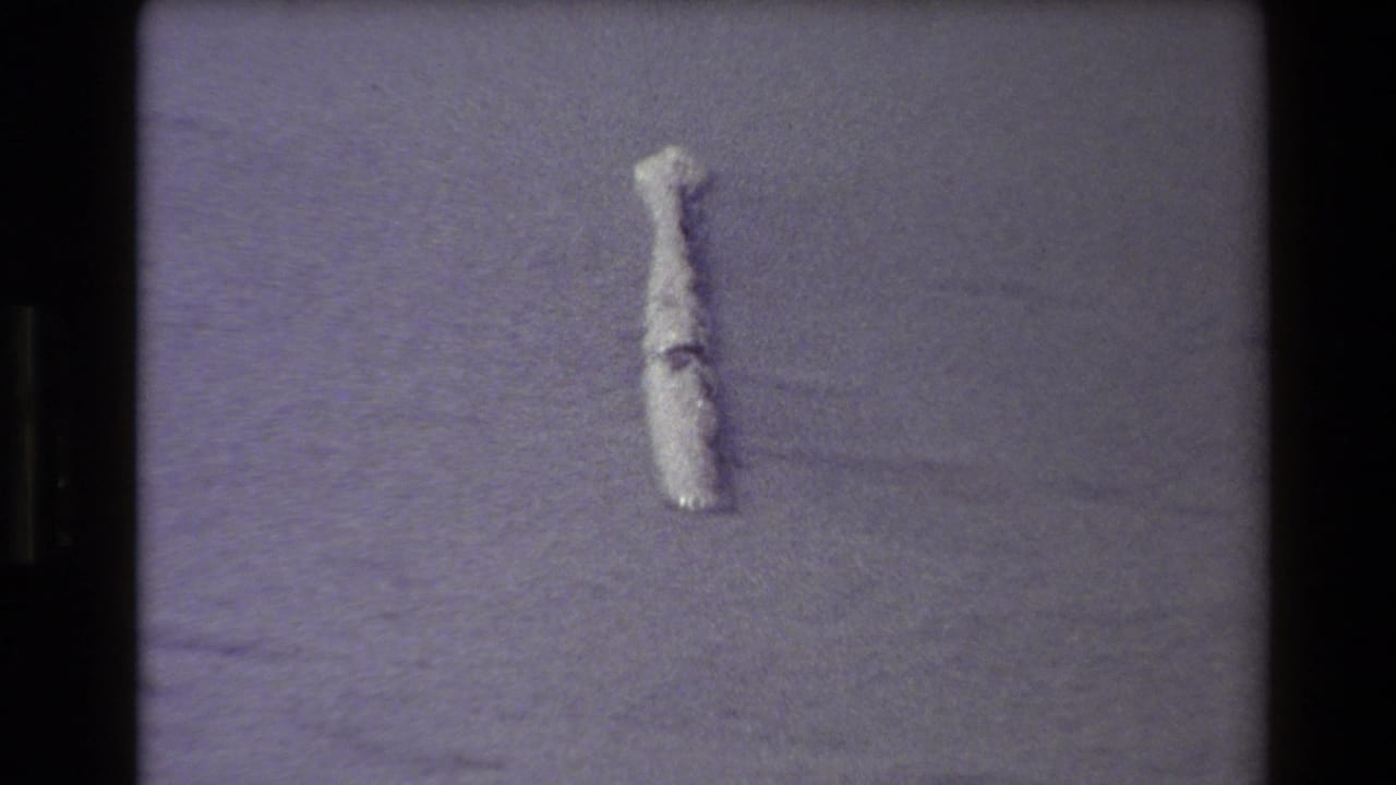1980 Western USA 8mm frame with a small pale cylindrical object on open sand, rendered with coarse grain and a cool purple cast.