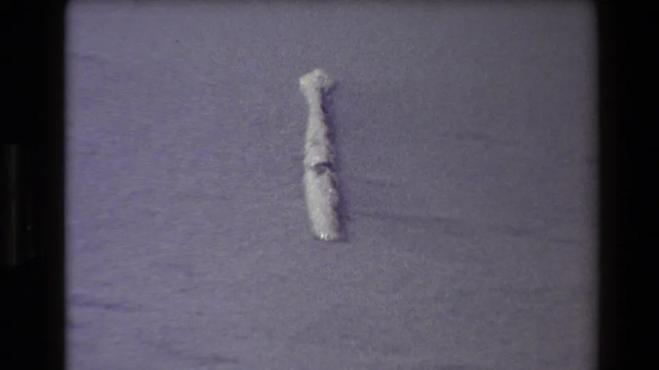 1980 Western USA 8mm frame with a small pale cylindrical object on open sand, rendered with coarse grain and a cool purple cast.
