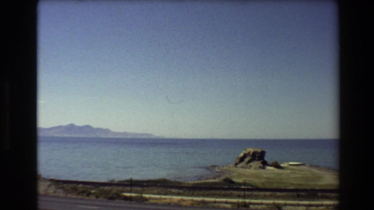 1980 Western USA 8mm frame showing blue sea, distant mountains, a rocky shoreline outcrop, and a roadside foreground from a moving vehicle.